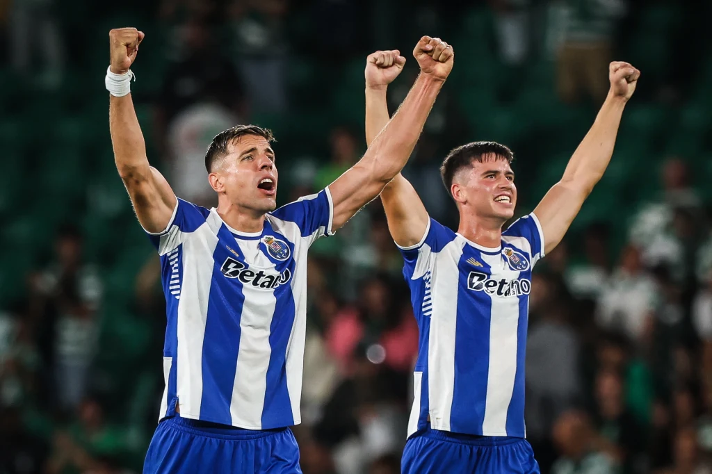 Jan Bednarek i Dominik Prpić z FC Porto Dwóch piłkarzy w niebiesko-białych koszulkach podnosi triumfalnie ręce, wykazując radość po zwycięstwie. W tle widoczny rozmazany tłum kibiców.