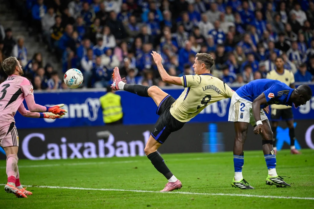 Robert Lewandowski na obiekcie Realu Oviedo Piłkarz w beżowo-czarnej koszulce wykonuje ekwilibrystyczny strzał do bramki podczas meczu, bramkarz w różowym stroju przygotowuje się do obrony, a obrońca w niebiesko-białym stroju odwraca się do akcji, wokół widoczna publiczność na trybunach.