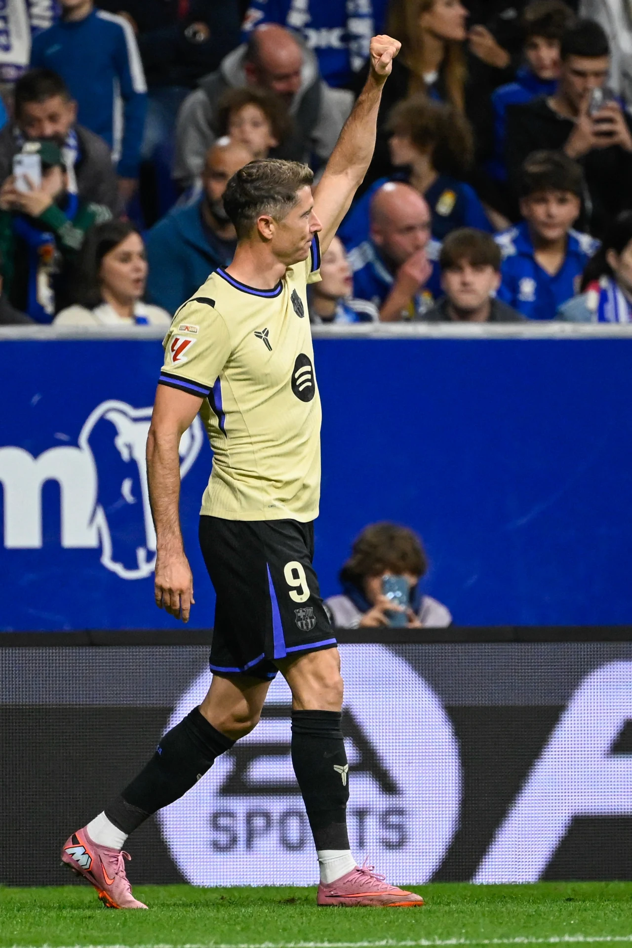 Robert Lewandowski tuż pod zdobyciu bramki Piłkarz w żółtej koszulce z numerem 9 unosi prawą rękę z zaciśniętą pięścią na tle trybun wypełnionych kibicami, w tle widać ludzi skupionych na wydarzeniu sportowym.