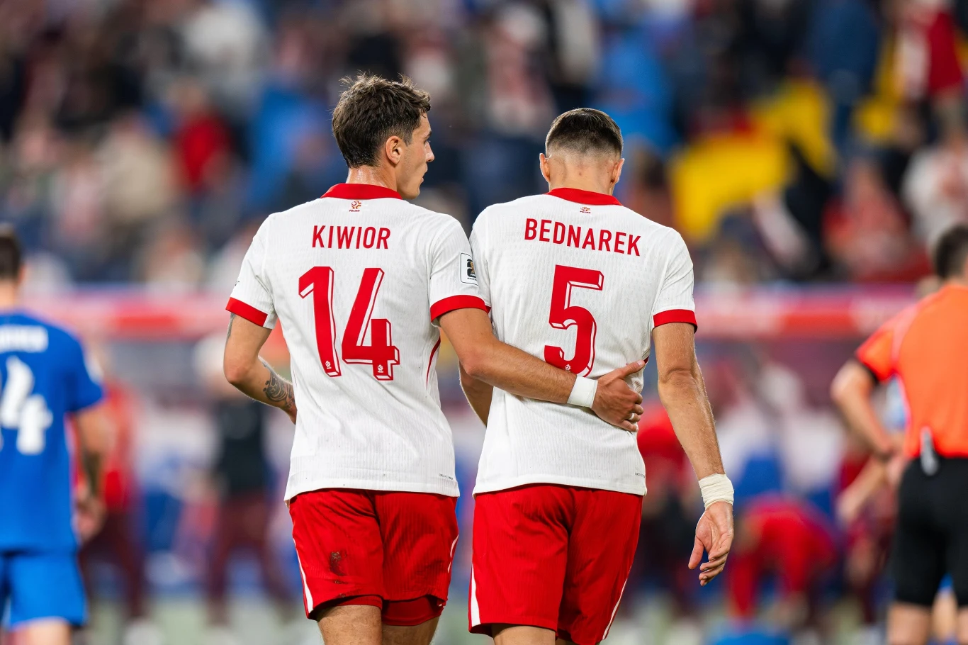 Jakub Kiwior, Jan Bednarek Dwóch piłkarzy drużyny w białych koszulkach z czerwonymi numerami idzie ramię w ramię po boisku, wokół nich rozmyci zawodnicy oraz kibice na trybunach.