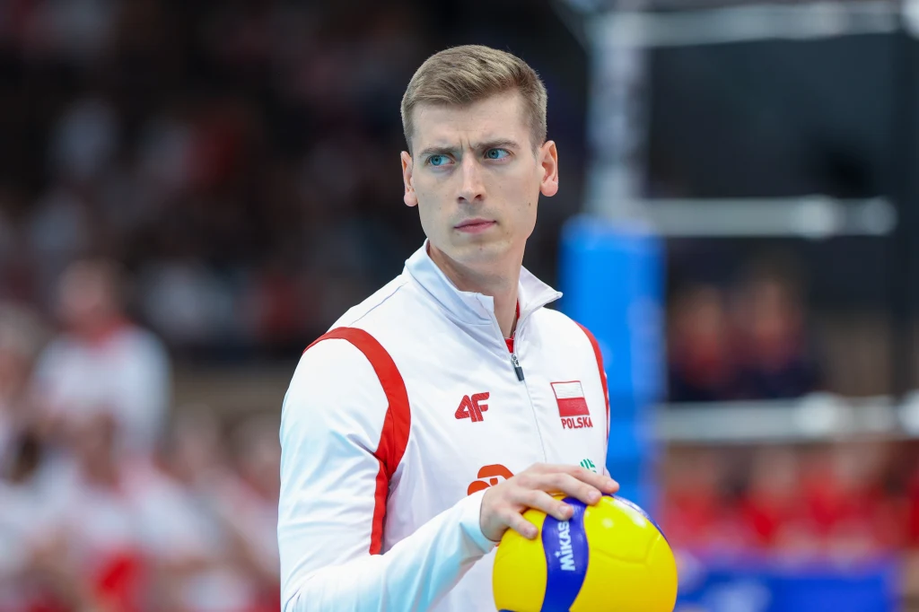 Kamil Semeniuk Siatkarz ubrany w biało-czerwony dres reprezentacji Polski trzymający piłkę do siatkówki na tle rozmytej publiczności w hali sportowej.