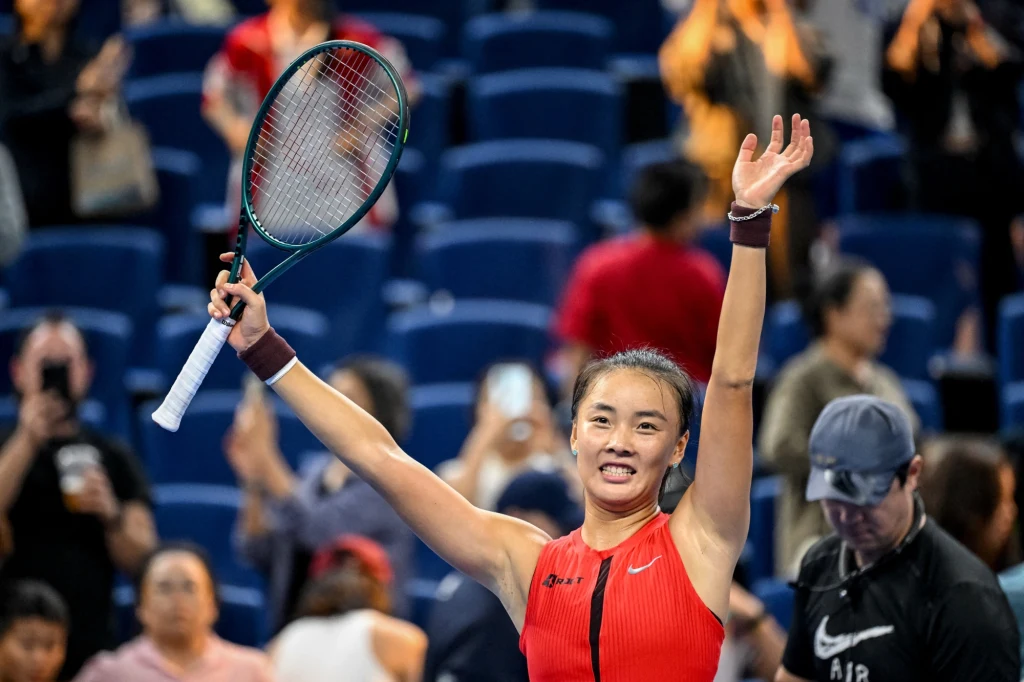 Yue Yuan Tenisistka w czerwonym stroju trzyma rakietę tenisową i unosi ręce w geście triumfu na korcie, w tle rozmyta publiczność klaszcząca i fotografująca moment zwycięstwa.