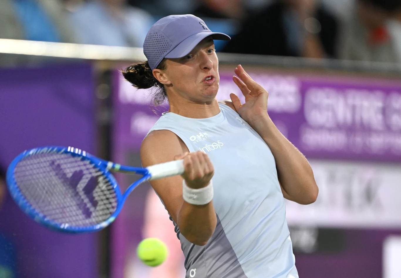 Tenisistka w jasnym stroju wykonuje forhend podczas meczu, dynamiczna akcja, widoczna piłka tenisowa i rakieta, w tle rozmazane trybuny.