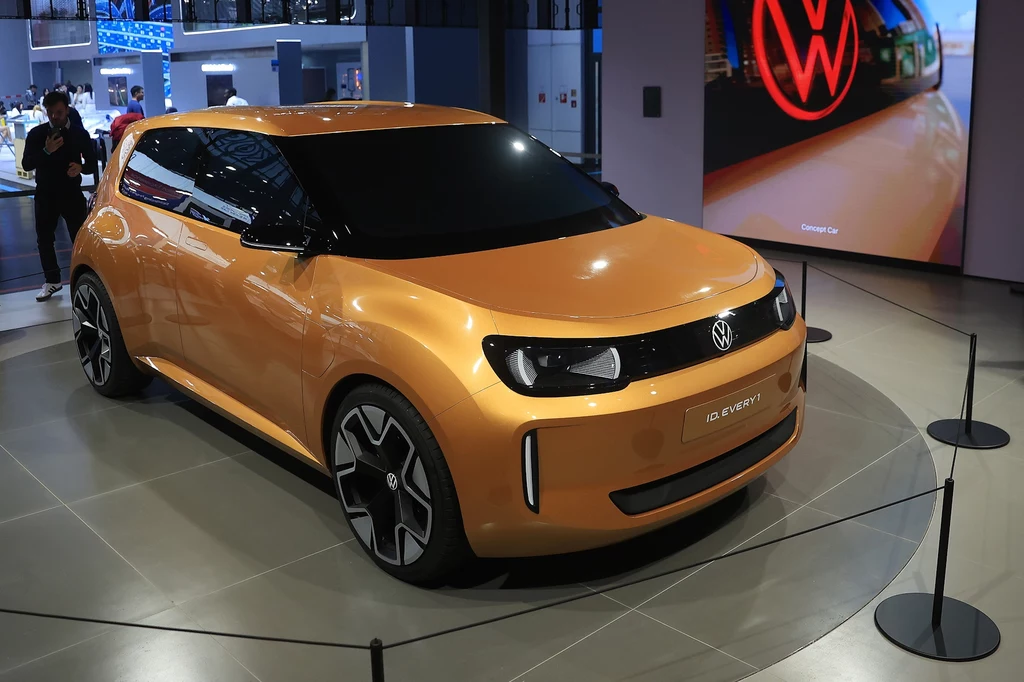 Pomarańczowy samochód koncepcyjny marki Volkswagen zaprezentowany na wystawie motoryzacyjnej, ustawiony na specjalnym podwyższeniu, w tle ekran z powiększonym logotypem producenta oraz sylwetki zwiedzających.