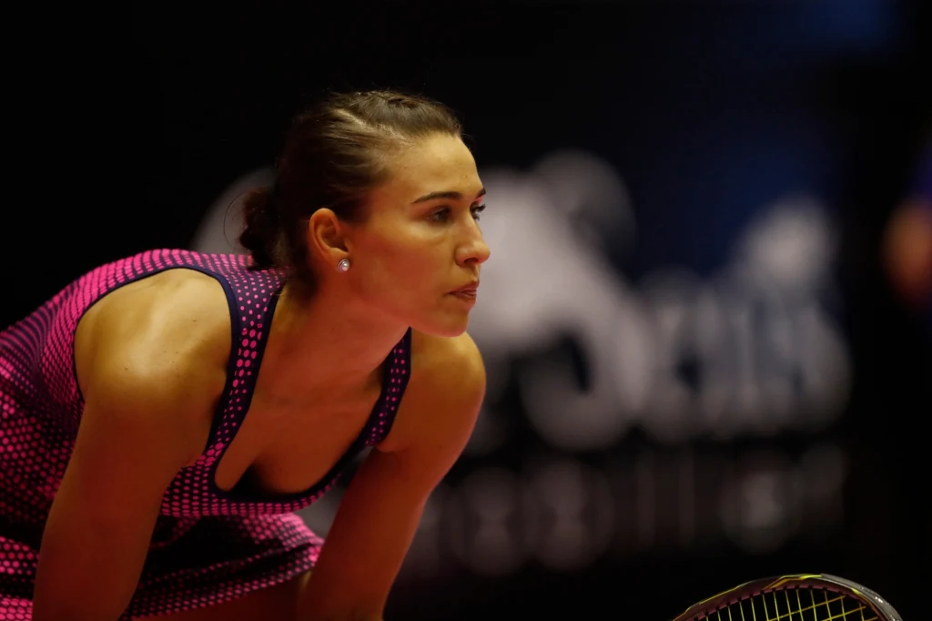 Witalija Djaczenko Kobieta w sportowym stroju tenisowym pochylona do przodu, skupiona, z rakietą tenisową w ręce na tle ciemnego, rozmytego tła z niewyraźnym napisem.