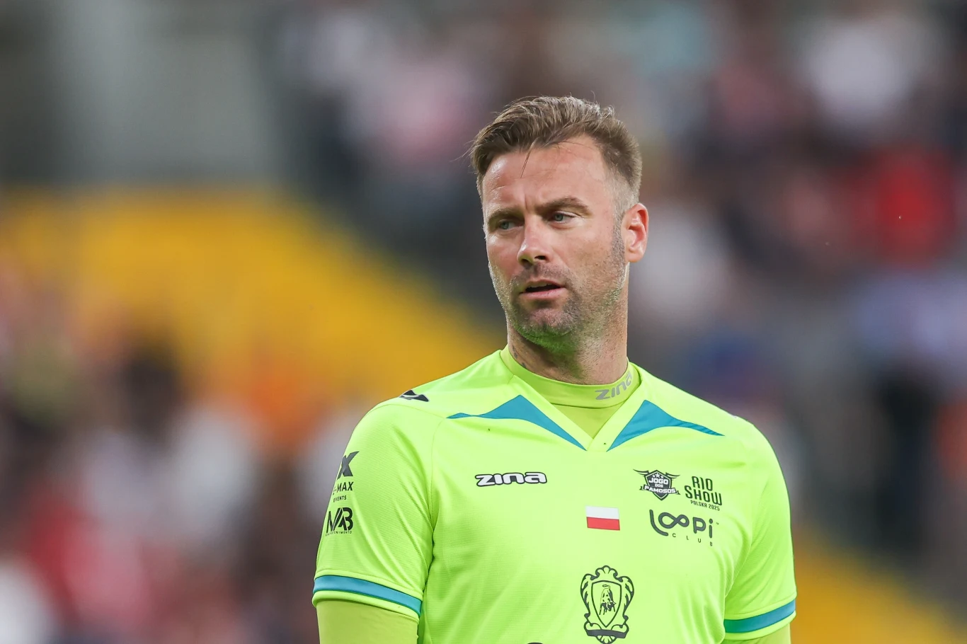 Artur Boruc Sportowiec w jasnozielonej koszulce sportowej z logotypami i flagą Polski na piersi, stojący na tle rozmytego stadionu z trybunami i widownią.