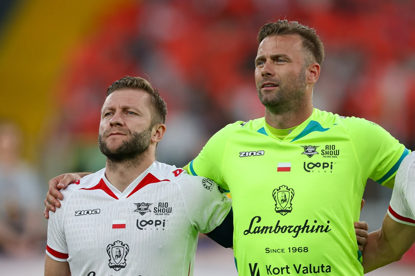 Artur Boruc Dwóch mężczyzn w strojach sportowych stoi obok siebie na boisku, obaj mają poważny wyraz twarzy, jeden ubrany w biały, drugi w jaskrawozielony strój piłkarski z widocznymi logotypami sponsorów.