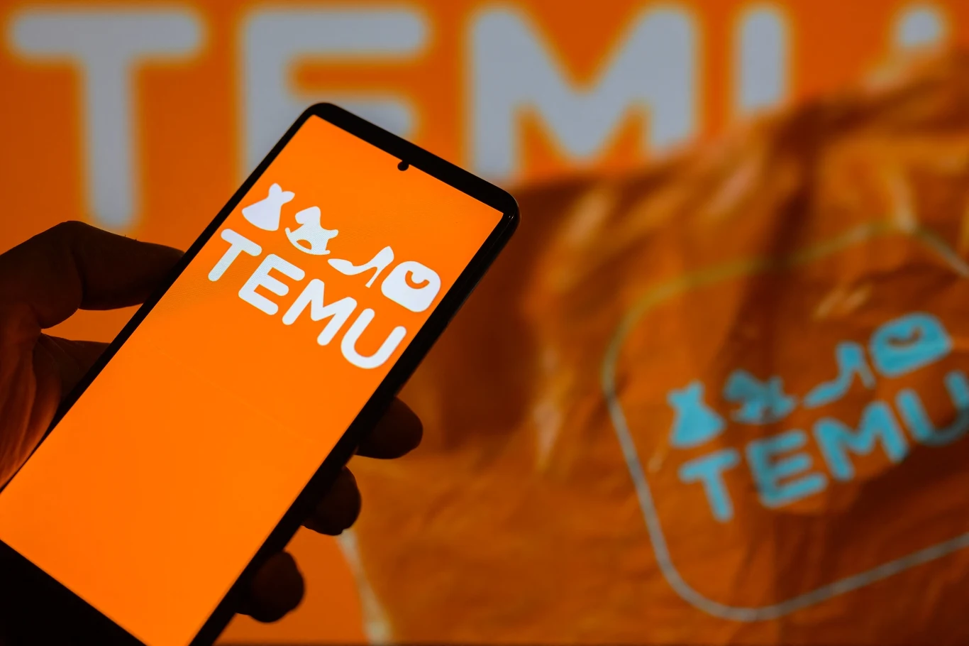 Zalew tanich paczek z Chin. UE przyspiesza wprowadzenie ceł w 2026 r. (zdj. ilustracyjne) Logo TEMU wyświetlone na ekranie smartfona trzymanego w dłoni na tle rozmazanego banera z logo TEMU w jednolitym, pomarańczowym kolorze.