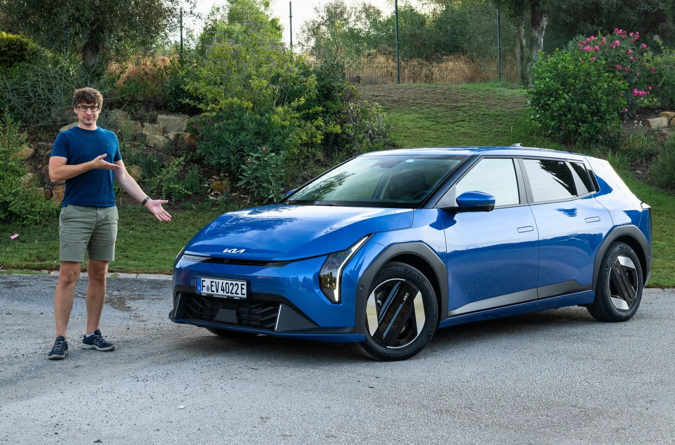 Kiedy Kia EV4 w Polsce? Mężczyzna w okularach i krótkich spodenkach stoi obok nowoczesnego, niebieskiego samochodu marki Kia, prezentując pojazd na tle zieleni i krzewów.