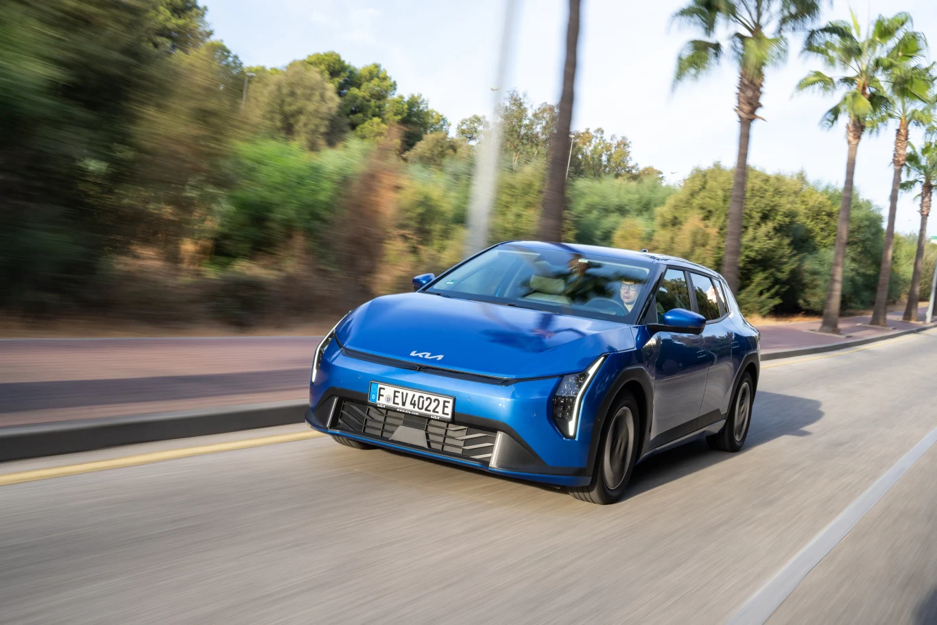 Kia EV4 z pewnością będzie się wyróżniać na drodze i z pewnością wzbudzi mieszane opinie; podobno tak miało być. Niebieski samochód elektryczny marki Aiways porusza się po pustej drodze, po bokach otoczony palmami i zielenią, w tle widoczne są drzewa i jasne niebo.