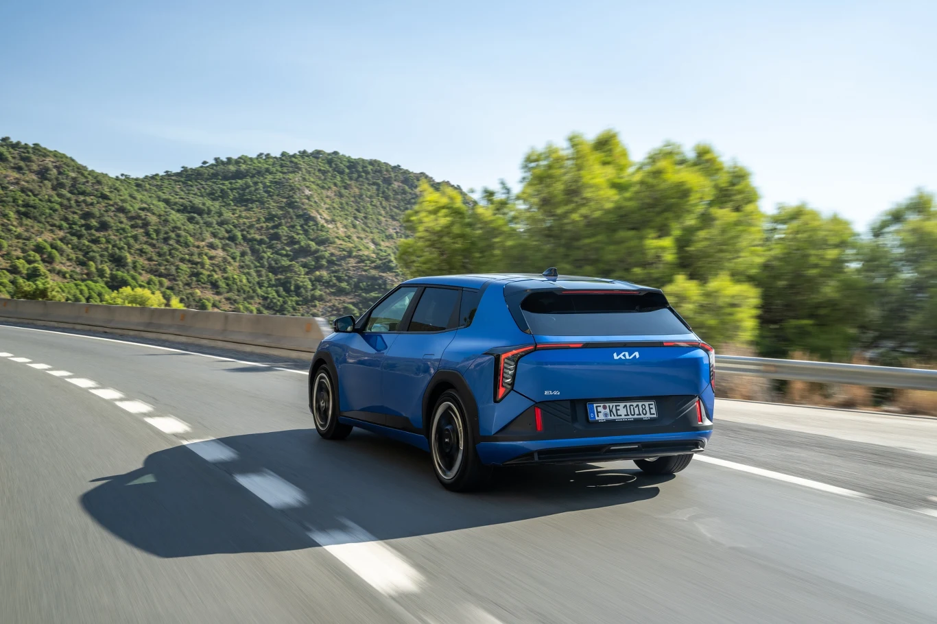 Charakterystyczne dla Kii są tylne lampy w kształcie bumerangów. Rozstaw osi jest imponujący, co przekłada się i na właściwości jezdne i na przestrzeń w kabinie. Niebieski samochód marki Kia jedzie dynamicznie po dobrze utrzymanej, krętej drodze otoczonej zielonymi wzgórzami i drzewami przy słonecznej pogodzie.