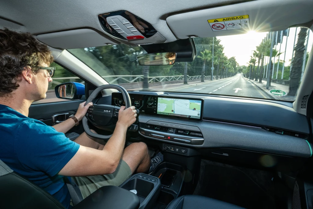 Młody mężczyzna prowadzi nowoczesny samochód elektryczny po pustej, szerokiej drodze, w kabinie wyraźnie widoczne są cyfrowe ekrany na desce rozdzielczej i jasne światło słoneczne przed pojazdem.
