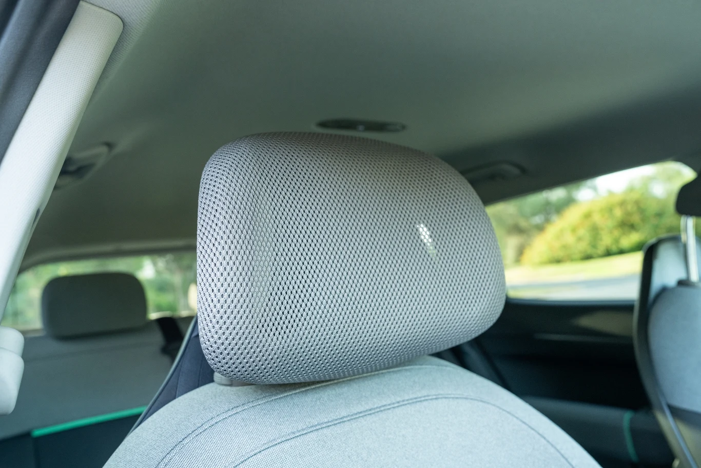 Zagłówki pokryte są prześwitującym materiałem typu “mesh”. Zbliżenie na zagłówek fotela samochodowego z jasnoszarego materiału, w tle widoczne wnętrze pojazdu oraz niewyraźny zarys okna i drzewa za szybą.