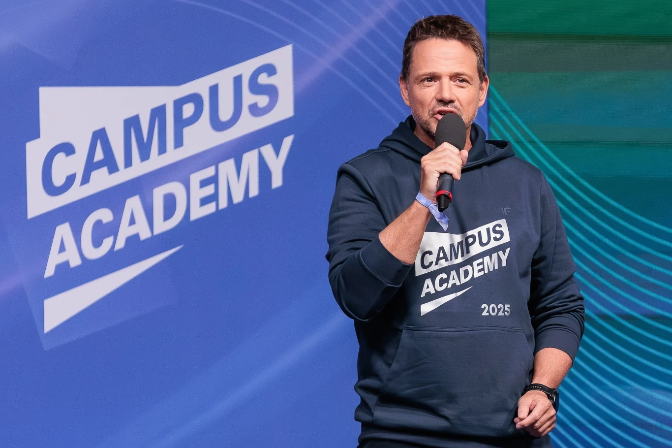 Mężczyzna w ciemnej bluzie z napisem Campus Academy 2025 przemawia do mikrofonu na tle niebieskiej planszy z logo Campus Academy.