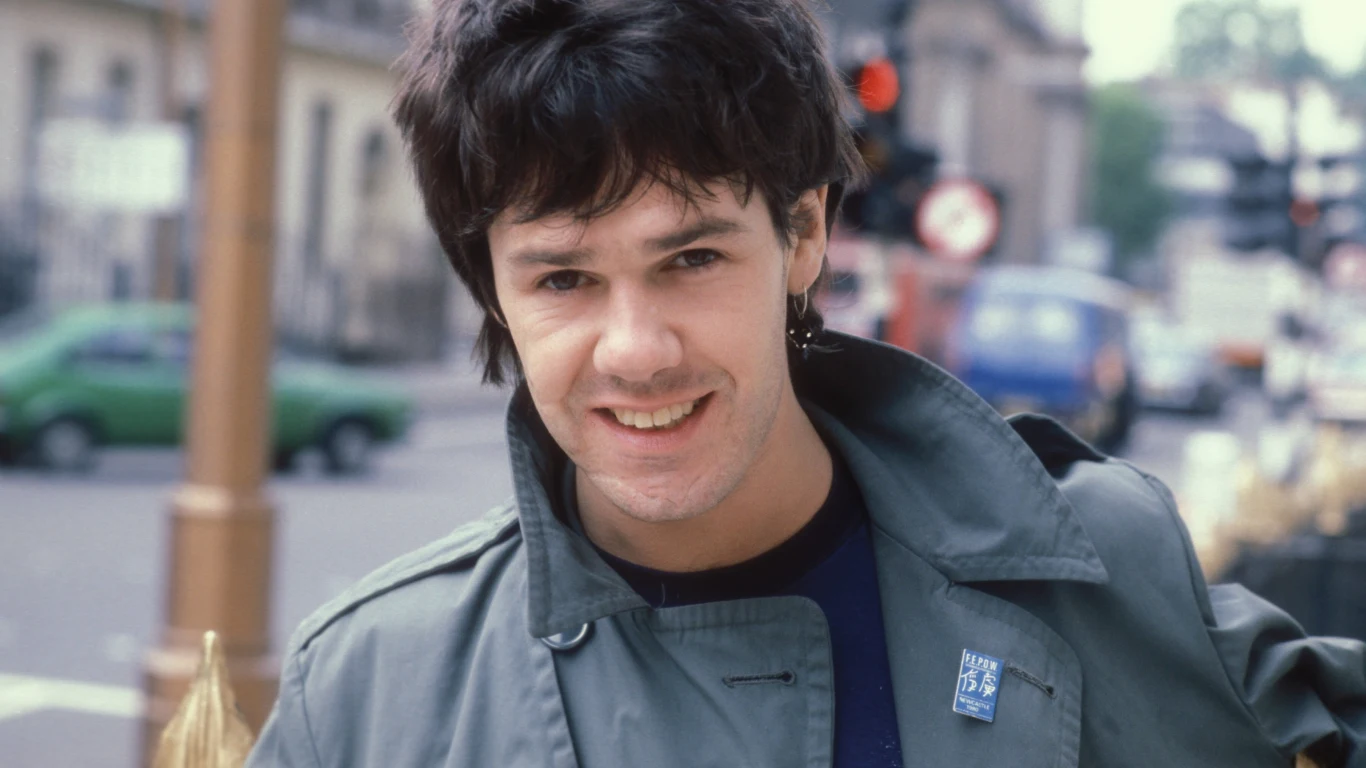 Gary Moore Uśmiechnięty młody mężczyzna z ciemnymi włosami ubrany w zieloną kurtkę, stojący na tle miejskiej ulicy z samochodami i rozmytym otoczeniem.