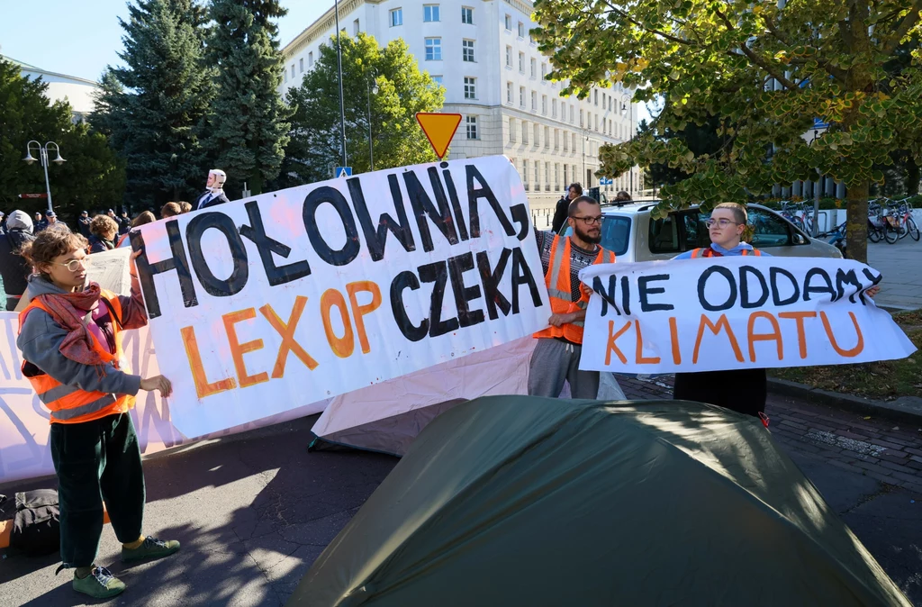 Grupa osób trzymających transparenty z napisami dotyczącymi ochrony klimatu i protestu przeciwko konkretnym rozwiązaniom politycznym, uczestnicy mają na sobie pomarańczowe kamizelki, w tle zielone drzewa i zabudowania miejskie