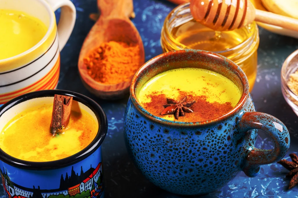 Zamień kawę na ten napój. Wzmacnia odporność i spowalnia starzenie Kubek z napojem o intensywnie żółtym kolorze, posypanym cynamonem i dekorowanym anyżem, obok stoją drugi kubek z cynamonem, słoik z miodem oraz drewniana łyżeczka z przyprawą kurkumą.