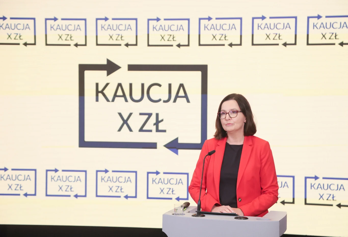 Kobieta w okularach i czerwonej marynarce stoi przy mównicy, w tle jasny ekran z napisem 'Kaucja X zł' oraz powtarzającymi się strzałkami i tym samym napisem.