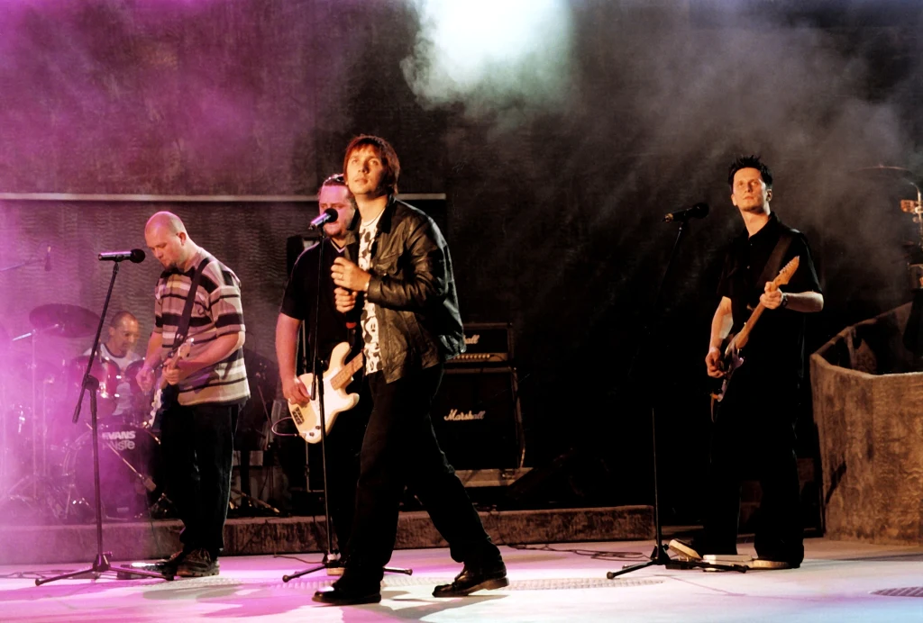 Zespół muzyczny wykonujący utwór na scenie, wokalista z gitarą na pierwszym planie, gitarzysta basowy i perkusista w tle, dynamiczne oświetlenie i atmosfera koncertu, fioletowe światła i dym podkreślają klimat wydarzenia.