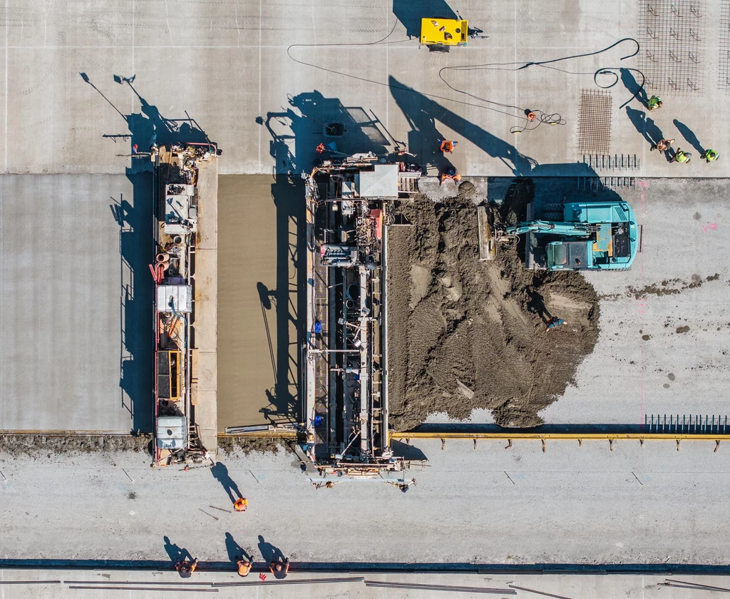 Ciężki sprzęt budowlany, w tym koparka i dwa duże urządzenia do wyrównywania nawierzchni, pracują przy układaniu i wygładzaniu betonu na dużej powierzchni drogi, kilku pracowników w kamizelkach odblaskowych nadzoruje działania z różnych stron placu budowy