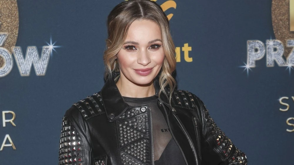 Ewelina Lisowska Kobieta ubrana w czarną skórzaną kurtkę z ćwiekami pozuje na tle niebieskiej ścianki z napisami, ma upięte włosy i delikatny makijaż