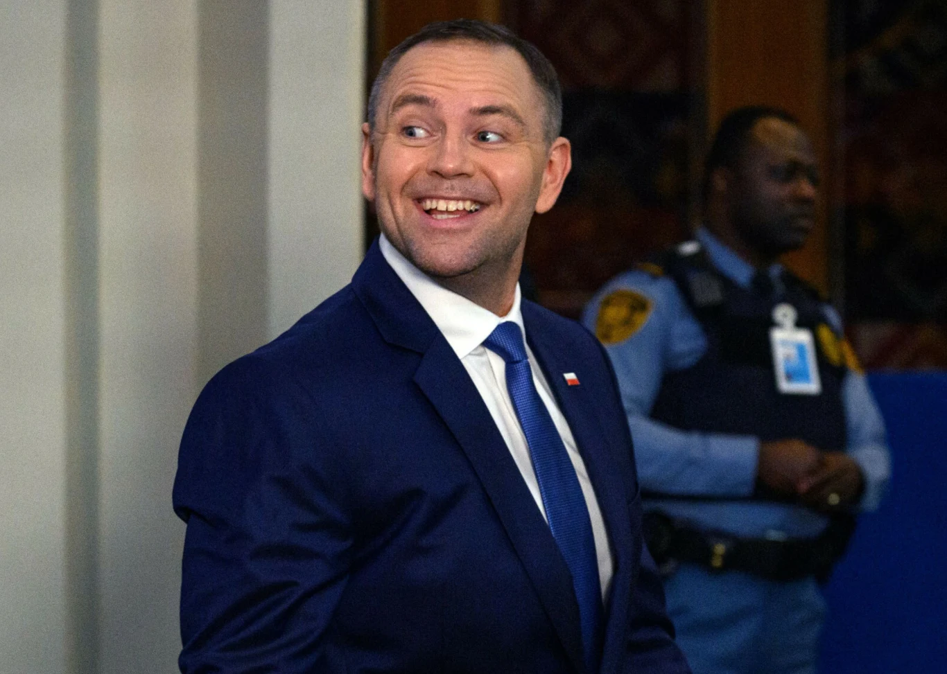 Prezydent RP Karol Nawrocki Mężczyzna w garniturze i pod krawatem uśmiecha się, stojąc wewnątrz budynku, w tle widoczny jest umundurowany ochroniarz.