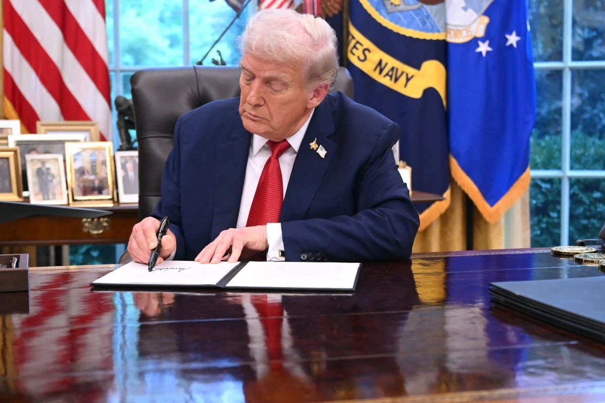 Mimo obietnic Trumpa klasa średnia traci wiarę w dobrobyt Donald Trump siedzi przy biurku w gabinecie owalnym, podpisując dokument, w tle widoczne są flagi oraz zdjęcia rodzinne.