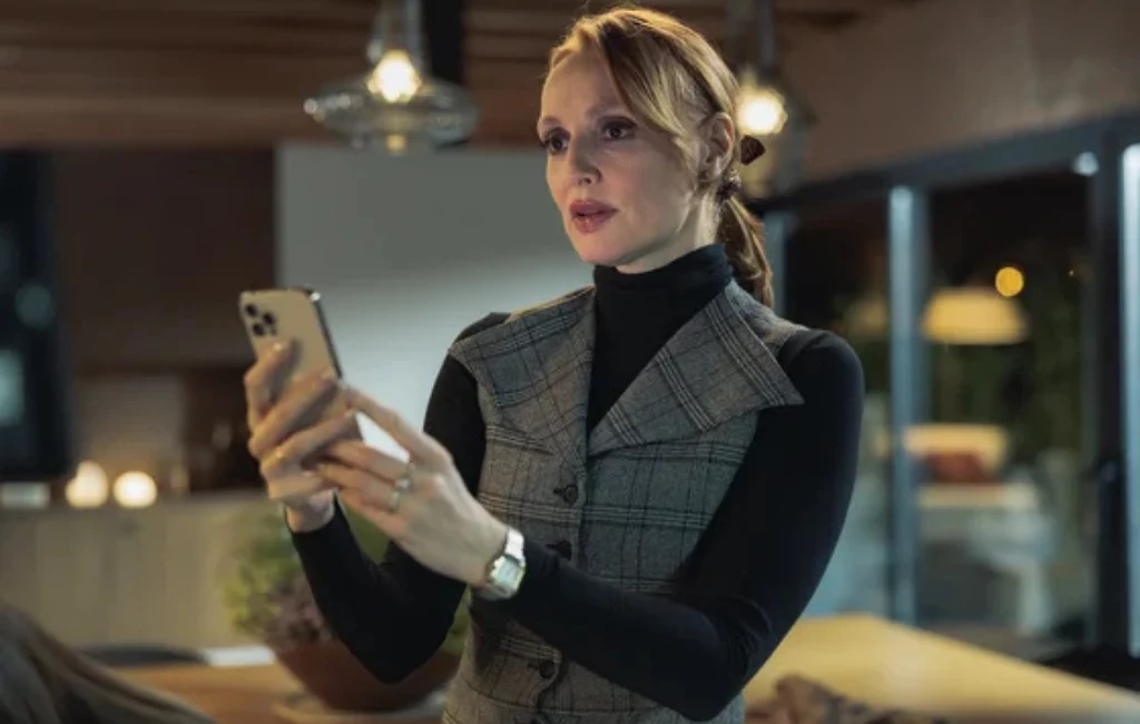 Kobieta z rudymi włosami ubrana w elegancką kamizelkę i czarny golf trzyma w dłoniach smartfon, znajduje się w nowoczesnym, przytulnie oświetlonym wnętrzu.