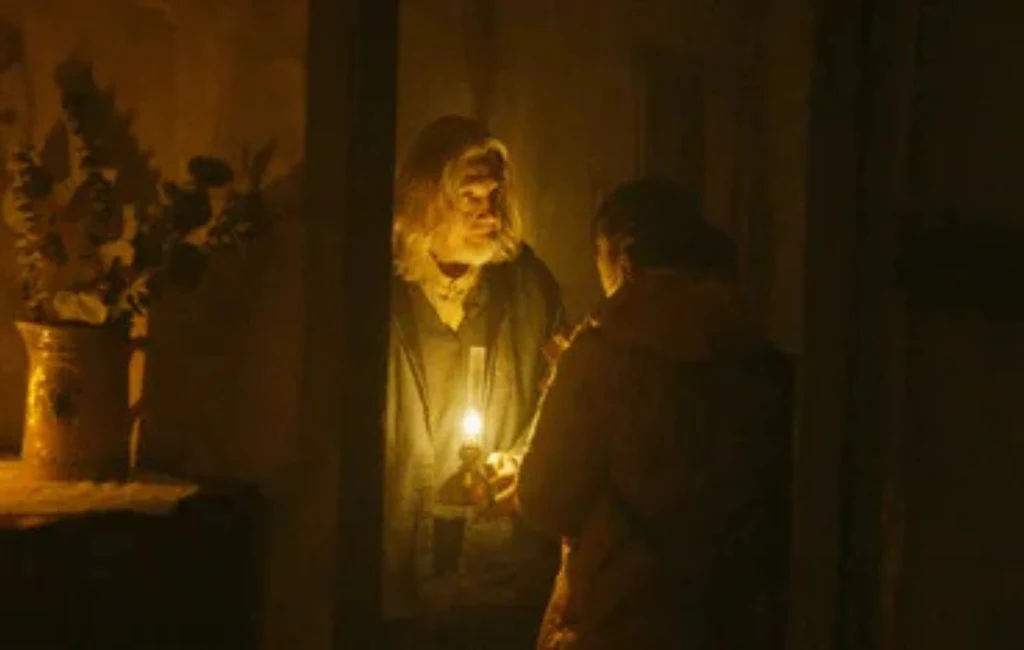 W rolach głównych: Emily Bader, Roland Buck III, Dan Lippert. Dwie osoby stoją naprzeciwko siebie w słabo oświetlonym pomieszczeniu, jedna z nich trzyma świecę, która rzuca ciepłe światło na ich twarze, w tle widoczna stara donica z kwiatami na szafce.