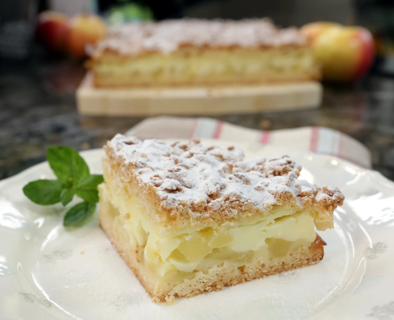 Szarlotka z budyniem. Przepis Ewy Wachowicz Kawałek szarlotki z kruszonką, nadzieniem z jabłek i kremem budyniowym, oprószony cukrem pudrem, na talerzu z listkiem mięty, w tle cały placek oraz jabłka.