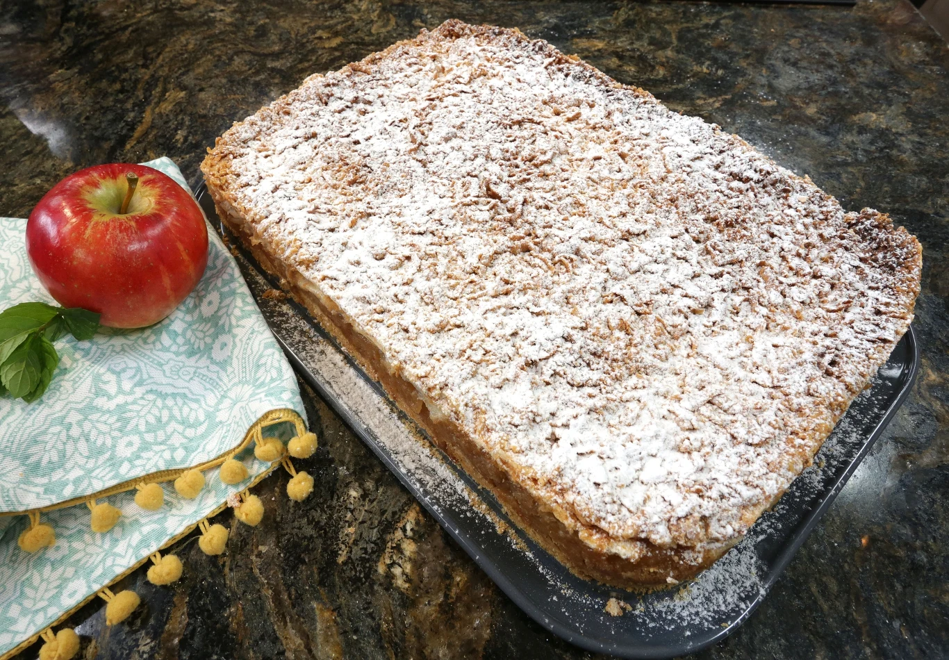 Klasyczna szarlotka. Przepis Ewy Wachowicz Duża prostokątna szarlotka oprószona cukrem pudrem, ułożona na ciemnym blacie kuchennym obok czerwonego jabłka, kilku listków mięty oraz ozdobnej ściereczki.