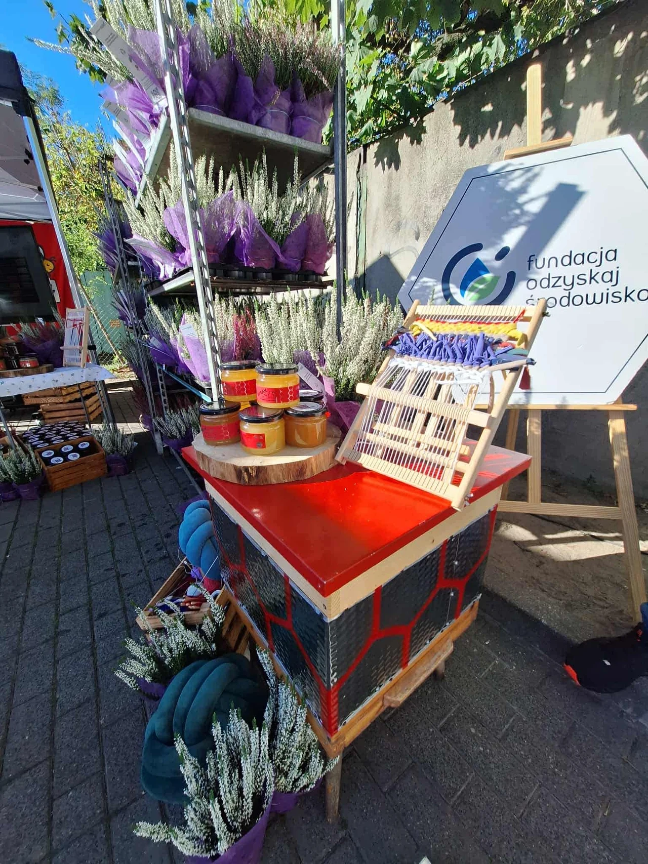 Stoisko z produktami pszczelarskimi zawierające kolorowy ul pokazowy, słoiki z miodem, rośliny lawendowe oraz elementy promocyjne fundacji związanej z ochroną środowiska. Tło stanowi regał z fioletowymi kwiatami, a z boku widoczny jest baner fundacyjny.