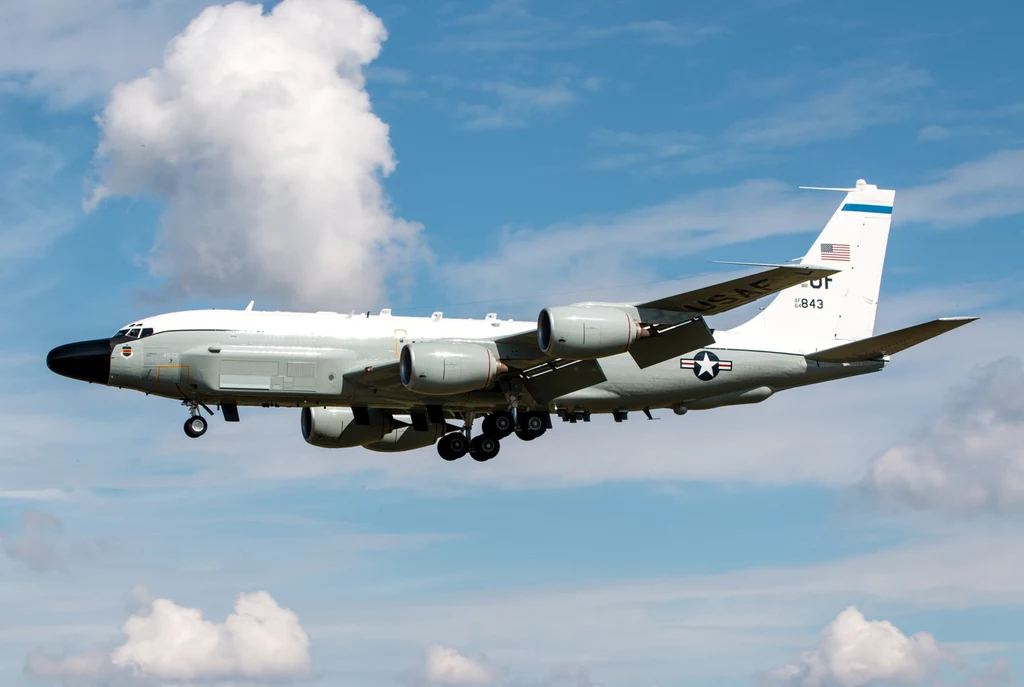 Wojskowy samolot zwiadowczy USAF RC-135 lecący na niewielkiej wysokości, wszystkie szczegóły samolotu widoczne są wyraźnie na tle niebieskiego nieba z pojedynczymi chmurami.