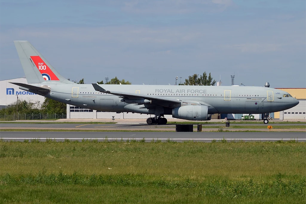 Duży wojskowy samolot transportowy oznakowany jako Royal Air Force stoi na pasie startowym, w tle widoczne są budynki lotniskowe i zieleń.