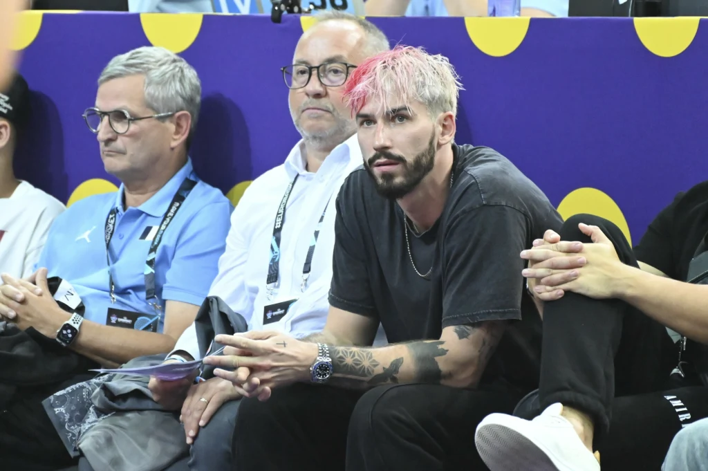 Mężczyzna z różowymi włosami i brodą siedzi na trybunach obok dwóch innych mężczyzn, jeden z nich ubrany w jasnoniebieską koszulkę, drugi w białą koszulę, wszyscy patrzą przed siebie, w tle fioletowa plansza z żółtymi kropkami.