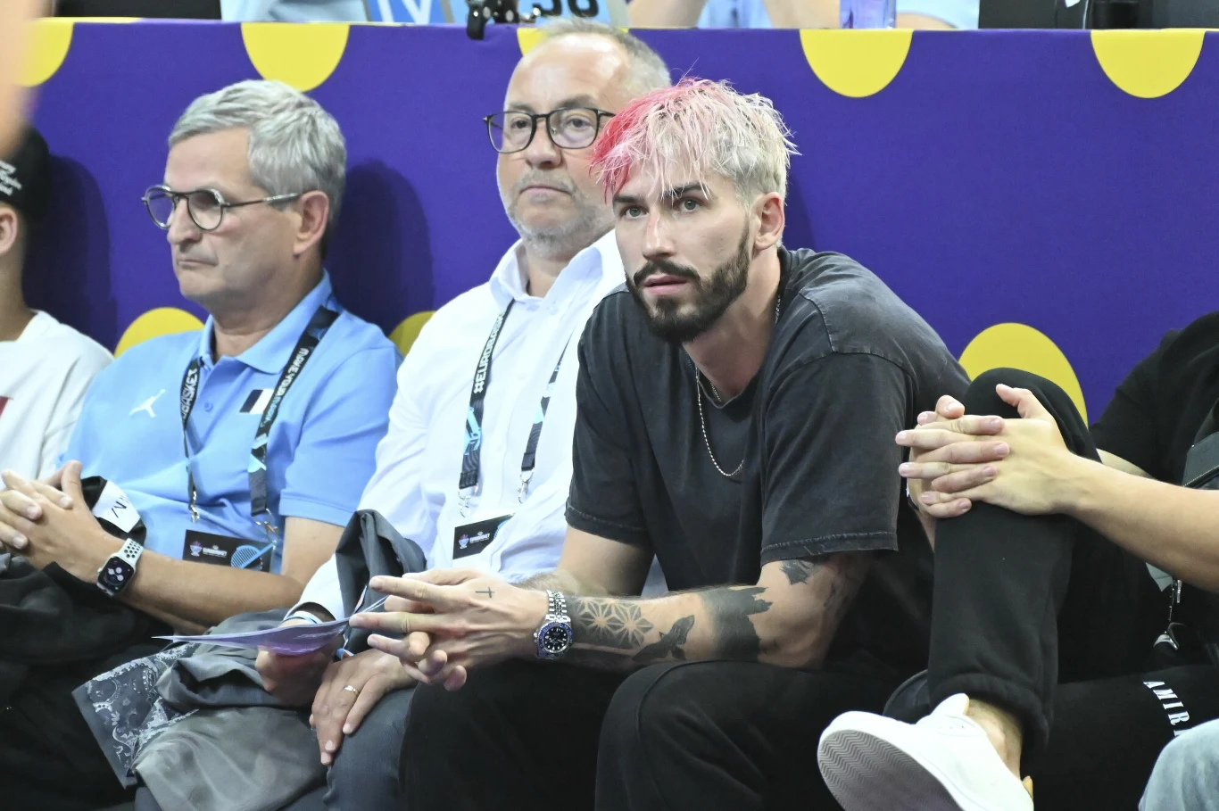 Mężczyzna z różowymi włosami i brodą siedzi na trybunach obok dwóch innych mężczyzn, jeden z nich ubrany w jasnoniebieską koszulkę, drugi w białą koszulę, wszyscy patrzą przed siebie, w tle fioletowa plansza z żółtymi kropkami.