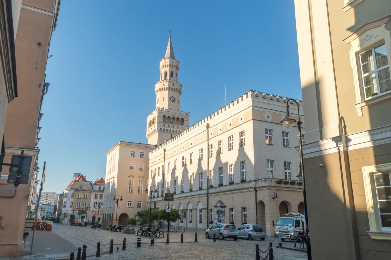 Opole to stolica województwa, gdzie zachwyca mnogość zabytków Zabytkowy budynek ratusza z wysoką wieżą zegarową, otoczony kilkupiętrowymi kamienicami, na brukowanej ulicy zaparkowane samochody, czyste niebo bez chmur.