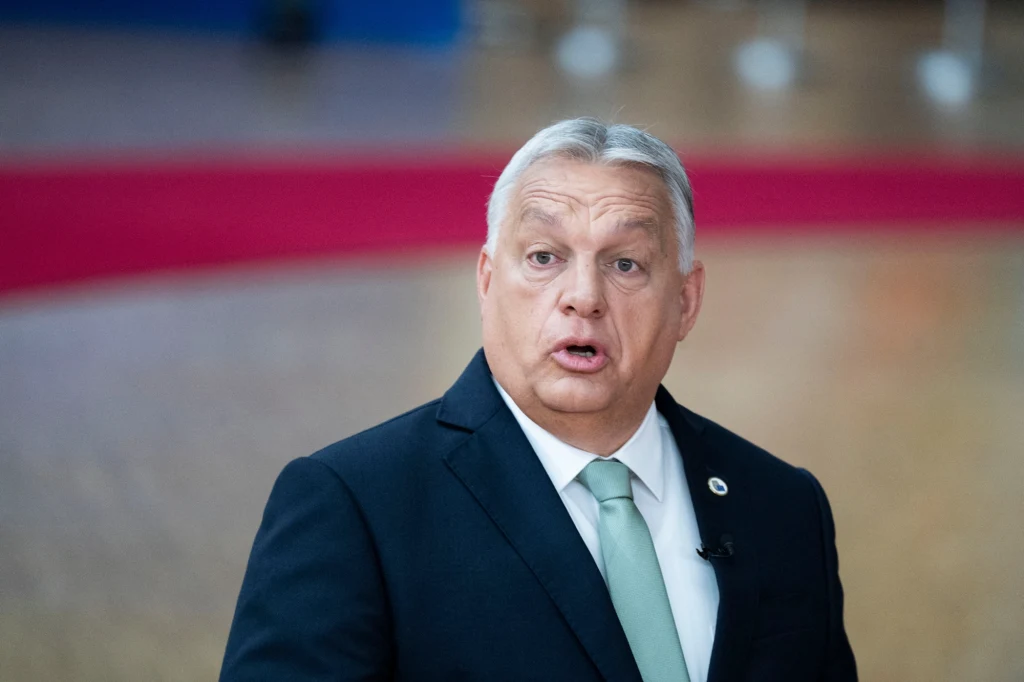 Mężczyzna w garniturze z jasnoniebieskim krawatem, patrzy z zaskoczeniem lub zdziwieniem, w tle rozmyte otoczenie z fragmentem czerwonego dywanu.