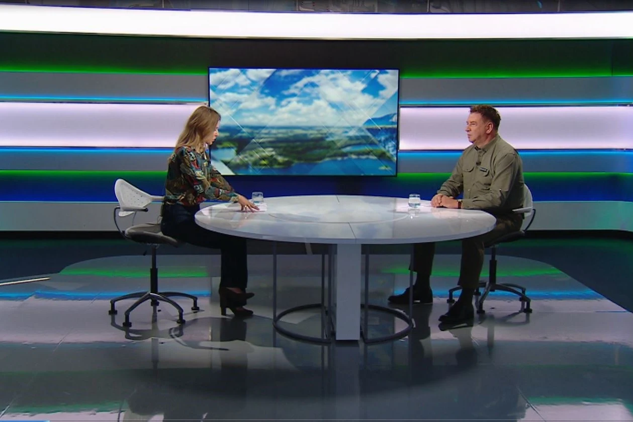 Rozmowa o ekologii na antenie Polsat News Dwóch dorosłych siedzących naprzeciwko siebie przy okrągłym stole podczas wywiadu telewizyjnego w nowoczesnym studiu z dużym ekranem w tle oraz szklankami wody na stole