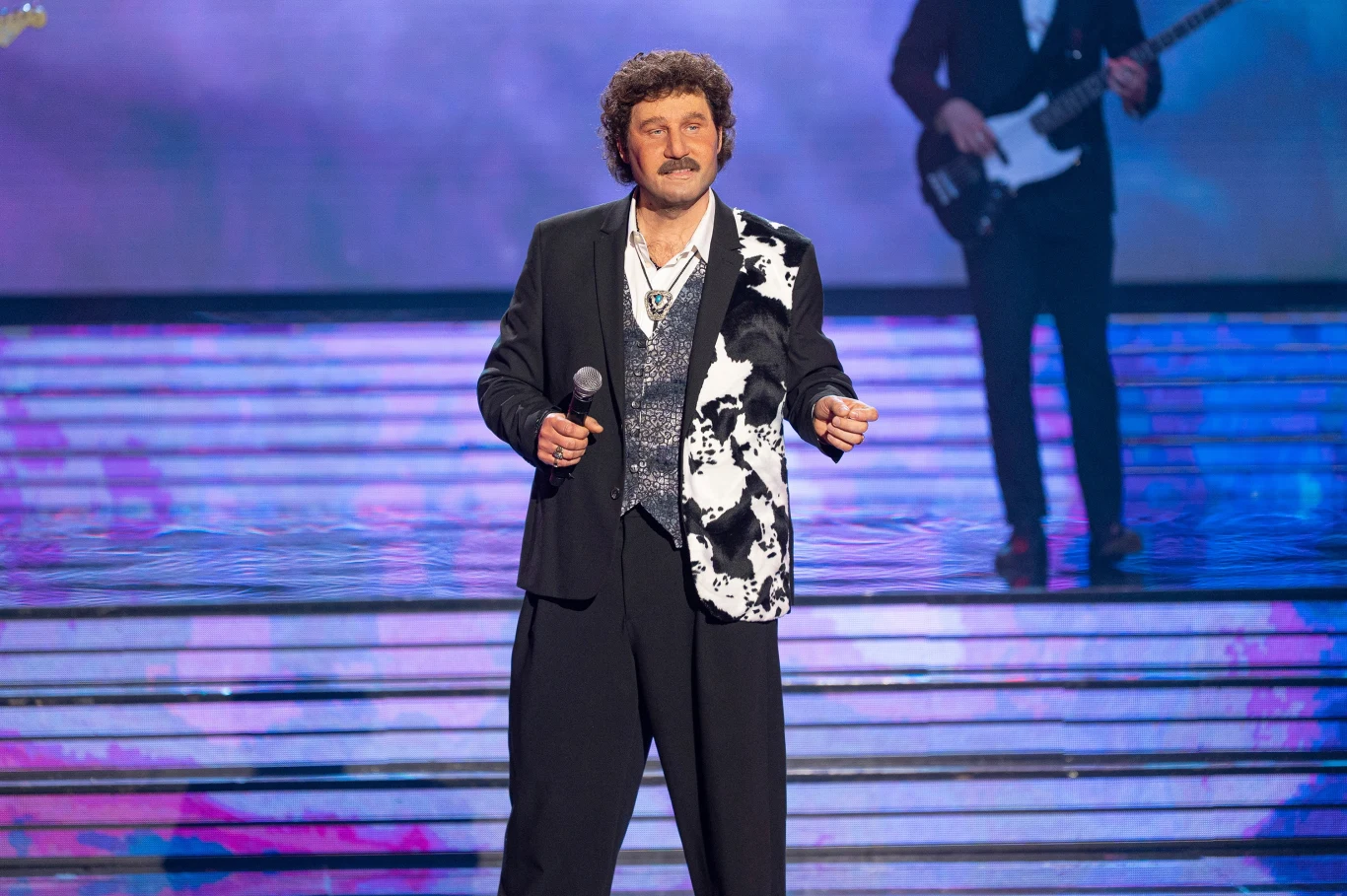 Andrzej Nejman jako Krzysztof Krawczyk Mężczyzna w eleganckim garniturze z mikrofonem stoi na scenie, w tle widoczny jest gitarzysta i kolorowo podświetlone schody.