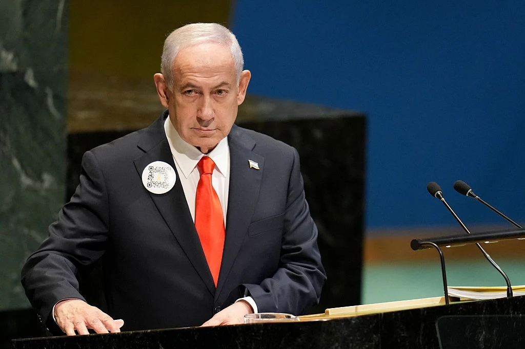 Mężczyzna w garniturze z czerwoną krawatą stoi za mównicą, w tle widoczny fragment sali konferencyjnej, na piersi przypięta okrągła plakietka.