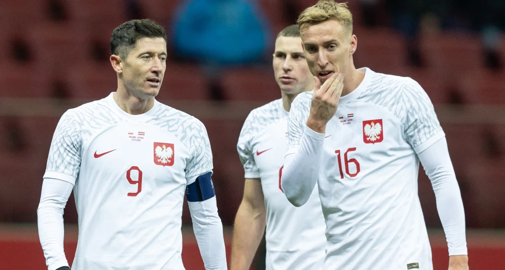 Adam Buksa (pierwszy z prawej) może nie otrzymać powołania na najbliższe mecze reprezentacji Polski Trzech piłkarzy polskiej reprezentacji w białych koszulkach z orłem na piersi podczas meczu, na pierwszym planie zawodnik z opaską kapitana i numerem 9 rozmawia z kolegą z drużyny, w tle trybuny stadionu.