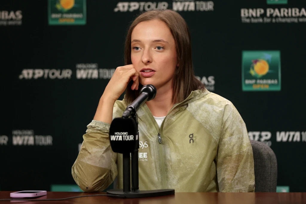 Kobieta siedząca przy stole podczas konferencji prasowej, trzymająca mikrofon, w tle logo WTA Tour oraz BNP Paribas, sportowe ubranie