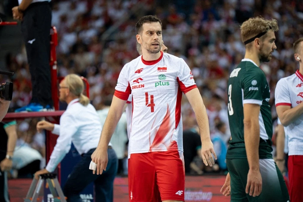 Siatkarze na boisku w trakcie meczu, pośrodku zawodnik w białej koszulce z czerwonymi akcentami i numerem 4, wokół niego widoczna publiczność oraz inni gracze w sportowych strojach