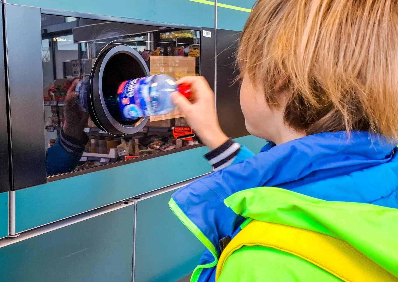 Do 3 listopada w Polsce zwrócono zaledwie 2300 puszek i butelek z kaucją Dziecko ubrana w kolorową kurtkę wrzuca plastikową butelkę do automatu do zbierania butelek, widoczny fragment sklepu w tle odbijający się w szybie maszyny.