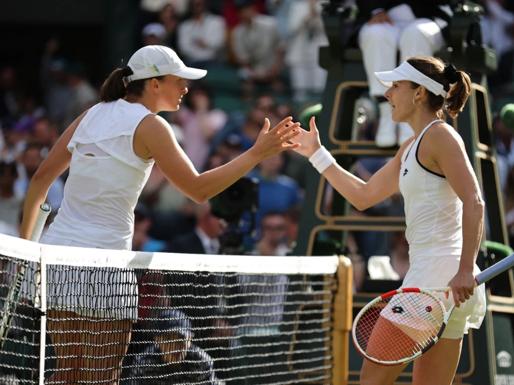 Dwie tenisistki w białych strojach na korcie podczas wymiany uścisku dłoni po meczu, widownia oraz elementy infrastruktury tenisowej w tle.