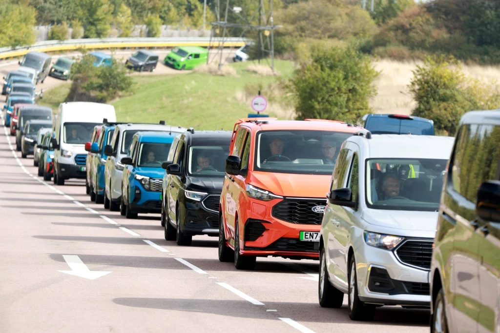 Szereg samochodów dostawczych różnych marek i kolorów ustawionych w kolejce na drodze, otoczonej zielenią i lekkim wzniesieniem z tyłu.