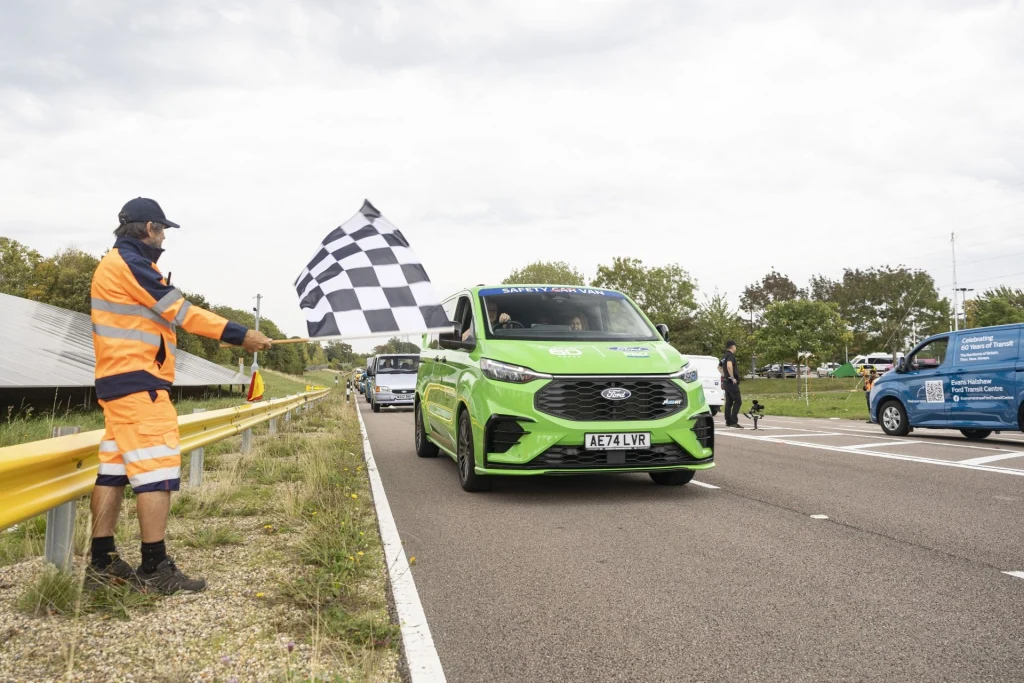 Zielony samochód przekracza linię mety na drodze, po lewej stronie osoba w kamizelce odblaskowej macha czarno-białą flagą w szachownicę, w tle widoczne inne pojazdy oraz fragmenty barier drogowych.