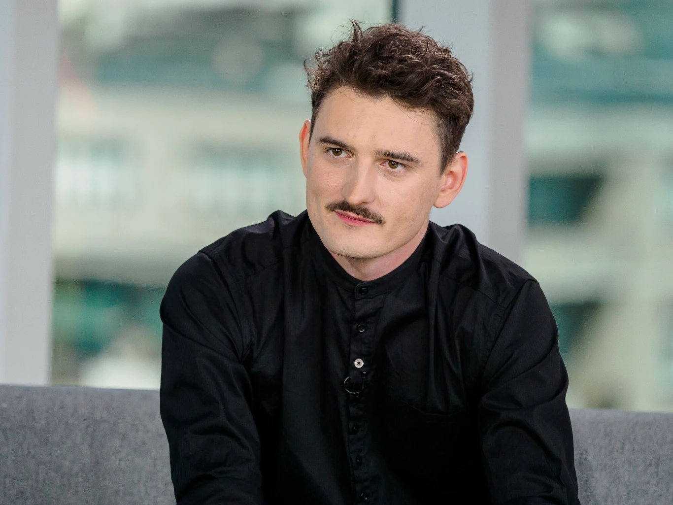 Dawid Podsiadło przygotował dla fanów niespodziankę Mężczyzna o ciemnych włosach i wąsach ubrany w czarną koszulę siedzi na szarej kanapie w nowoczesnym wnętrzu z dużym oknem w tle.