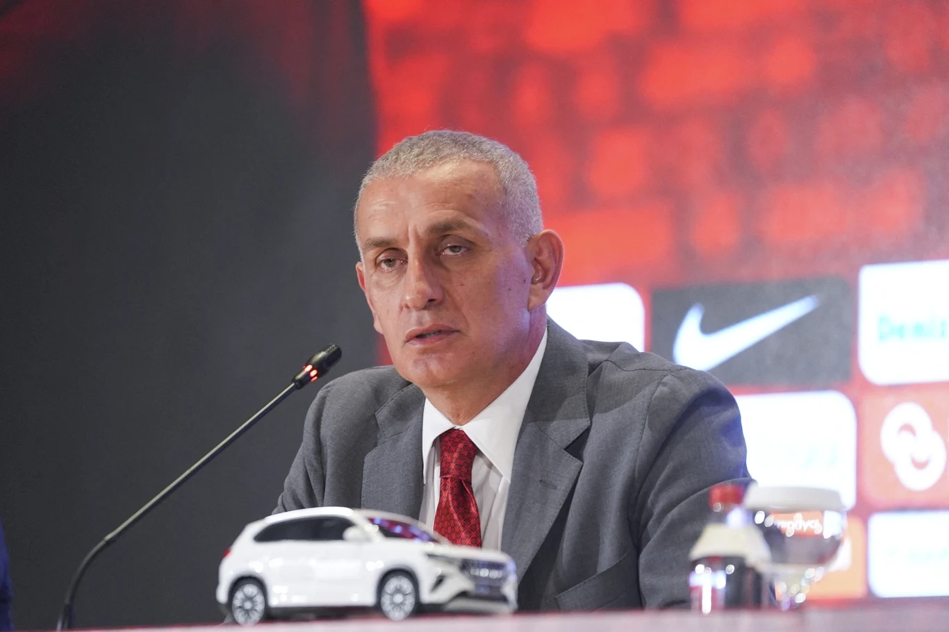 Mężczyzna w szarym garniturze i czerwonym krawacie siedzący przy stole konferencyjnym z mikrofonem, przed nim model samochodu i szklanka z wodą, w tle widoczne rozmazane logo Nike na czerwonym tle.